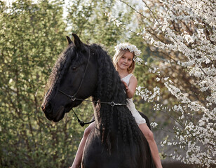 Beautiful little girl with white hair in a spring wreath riding a Friesian horse