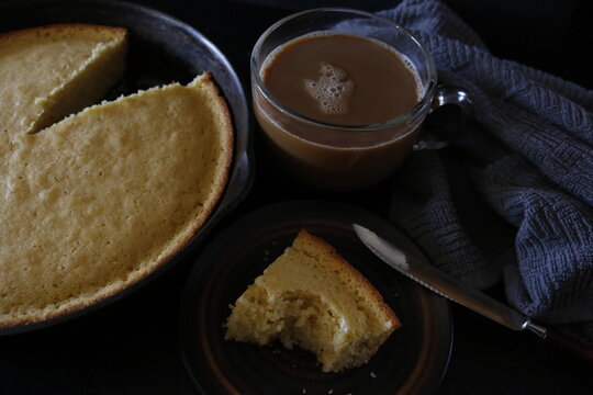 Studio shot of a cup of coffee with milk next to a cast iron skillet of freshly baked homemade Southern style cornbread with a piece cut out of it.