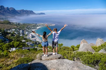 Acrylic prints Table Mountain Couple men and women with hands up standing on a mountain top The Rock viewpoint in Cape Town over Campsbay, with fog over the ocean. fog coming in from the ocean at Camps Bay Cape Town South Africa