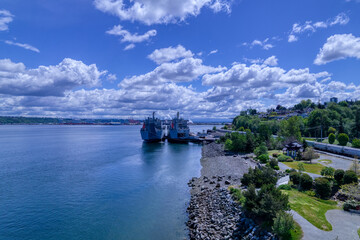 Two ships docked on the rocky shores of Puget Sound in Tacoma, Washington under a blue cloud filled...