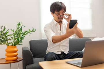 Middle age Asian man squint eye to look at the phone due to long eye sighted problems, which makes...