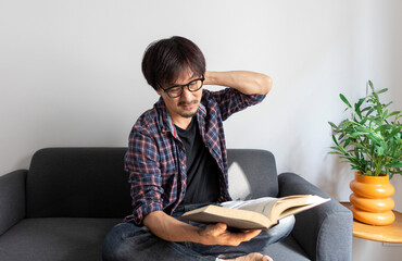 Asian man rubbing eye with tired expression after long period reading book, sitting on sofa at home.