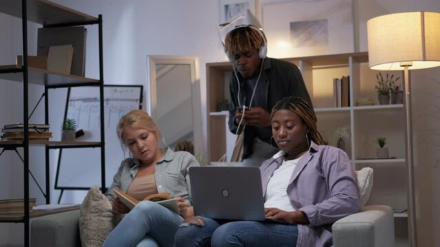 Students home leisure. Different lifestyle. Group hangout. Smart diverse girls learning using laptop reading book careless guy dancing in headphones in modern campus interior.