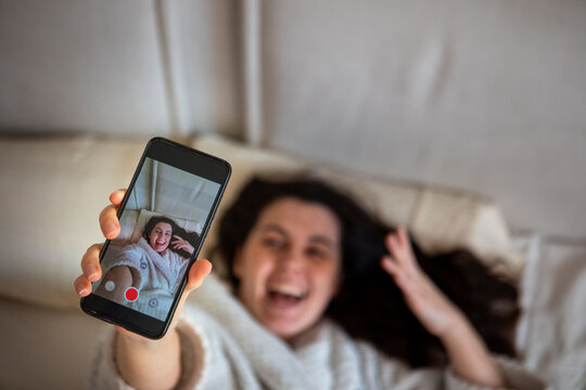 young happy woman taking selfie story laying down in bed