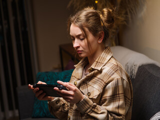 A fair-haired girl plays a video game on a console at home in a room in the evening. Recreation, relaxation, hobby, strategy games, virtual reality, games with friends online. Profile shot.