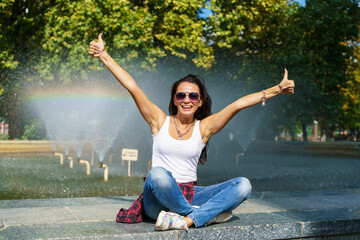 Fototapeta na wymiar Cute caucasian woman sits by fountain in park on sunny day, enjoys the coolness from the water, happy time spending on a day off