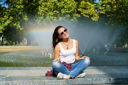 Cute caucasian woman sits by fountain in park on sunny day, enjoys the coolness from the water, happy time spending on a day off