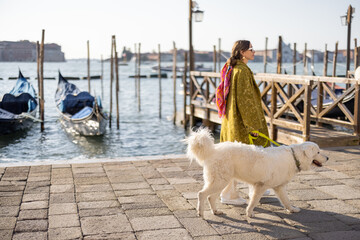 Young woman walking with dog near water canal with gondolas at saint Marks square in Venice. Woman wearing coat and colorful shawl in italian style. Maremmano abruzzese Italian sheepdog - obrazy, fototapety, plakaty
