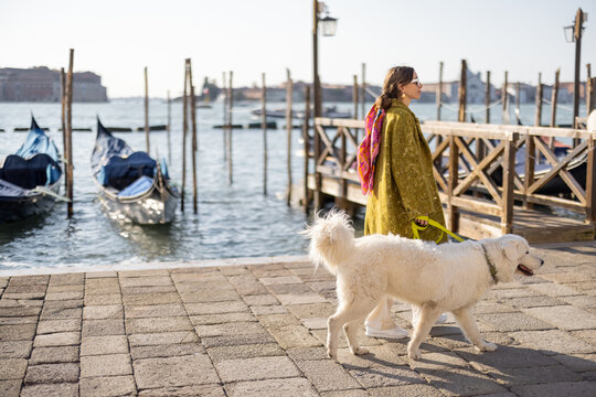 Young woman walking with dog near water canal with gondolas at saint Marks square in Venice. Woman wearing coat and colorful shawl in italian style. Maremmano abruzzese Italian sheepdog