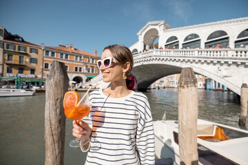 Young woman drinks summer cocktail on the background of famous Rialto bridge in Venice. Concept of happy vacation and leisure time in Italy. Holding italian alcohol drink spritz Aperol