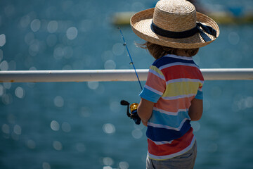 A young boy sits on the lake, dreams. Warm summer or spring day. Cute caucasian kid fishing. Little boy fishing in a pond view from behind.