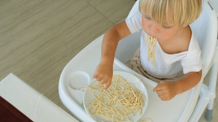 Baby boy eats spaghetti sitting in a baby chair in restaurant