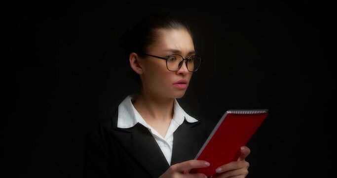 Female boss in glasses and office clothes is given red notebook from outside the frame.Business woman with a dissatisfied face reads a document,spreads her hands,looks at the camera.Black background