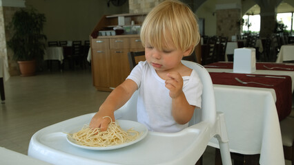 Baby boy eats spaghetti sitting in a baby chair in restaurant