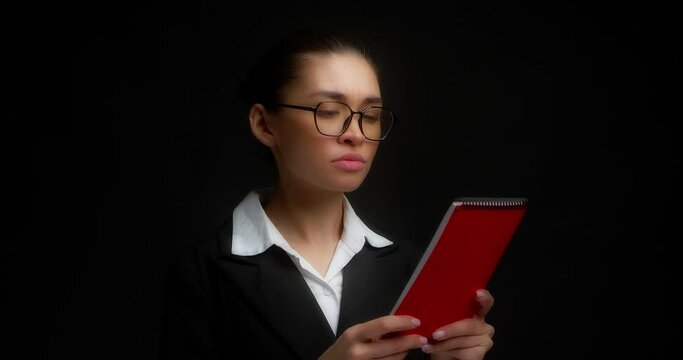 Female boss in glasses and office clothes is given red notebook from outside the frame.Business woman with a dissatisfied face reads a document,spreads her hands,looks at the manager.Black background