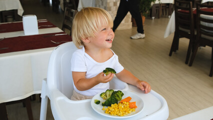 a little boy sits in a highchair and eats broccoli