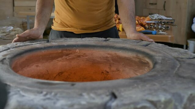 a male baker leans his hands on a stone tonir and looks inside as the fire burns inside the tandoor