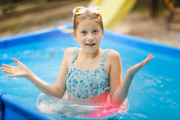 European preteen girl in sunglasses and swimsuit swims on circle in pool backyard. Funny child in pink glasses with shells and with an inflatable ring with pink feathers