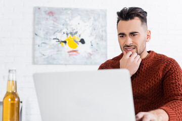 worried bookmaker looking at laptop near blurred bottle of beer.