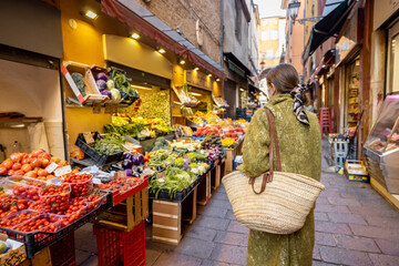 Woman in medical mask choosing fresh products at market stall on the famous gastronomical street in...