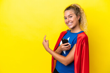 Super Hero woman isolated on yellow background using mobile phone and pointing back
