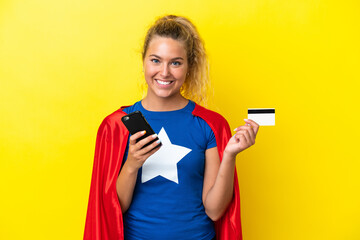 Super Hero woman isolated on yellow background buying with the mobile with a credit card