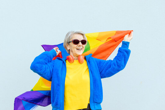 Happy 80 Years Old Woman Dressed Colorfully In Sunglasses Proudly Waving The Rainbow Flag Of The Gay Community