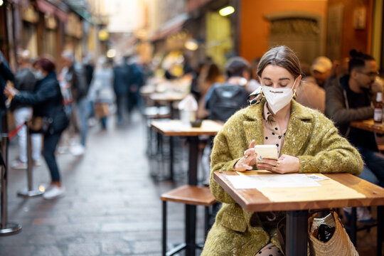Woman in medical mask sitting with phone at outdoor restaurant on crowded street in Bologna city. Concept of Italian gastronomic culture and new social rules related to pandemic