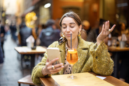 Young woman using phone while sitting with spritz aperol drink at bar outdoors. Concept of Italian lifestyle and gastronomy. Idea of remote online communication