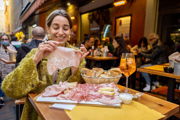 Woman having fun, while eating meat plate at outdoor bar in Bologna city. Girl holds mortadella,...