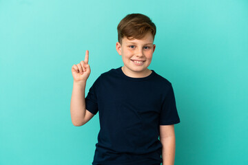 Little redhead boy isolated on blue background showing and lifting a finger in sign of the best