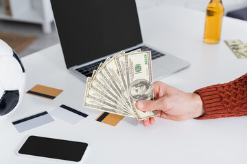 cropped view of bookmaker holding dollars near laptop, credit cards and soccer ball.