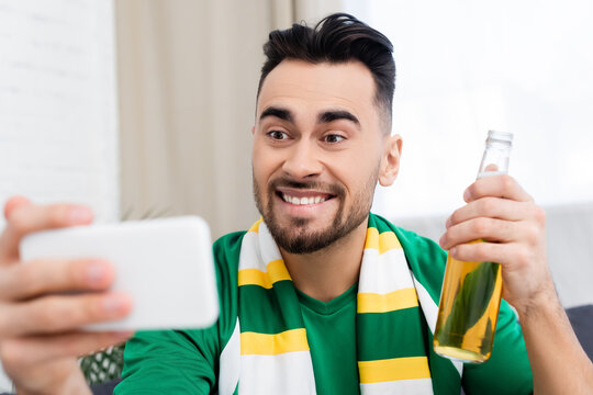 excited sports fan with bottle of beer taking selfie on blurred smartphone.