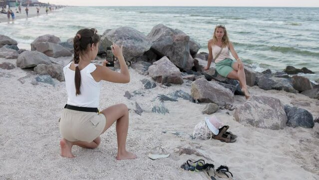 A girl with a scythe and a mobile phone takes pictures of her mother who is sitting on a stone on the sea coast