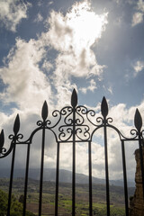 wrought iron fence with the sun behind white clouds with a blue sky and a town in the background