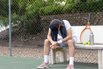 Exhausted sportsman sitting on a bench after tennis training class looking down to the floor in the...