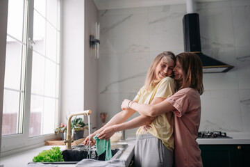 A lesbian couple is standing near the sink in the kitchen, washing dishes, hugging and laughing