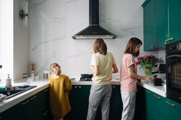 A lesbian family is standing in a green kitchen with their backs to the camera and preparing breakfast