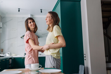 Lesbian couple is standing next to the dining table in the kitchen, hugging and laughing