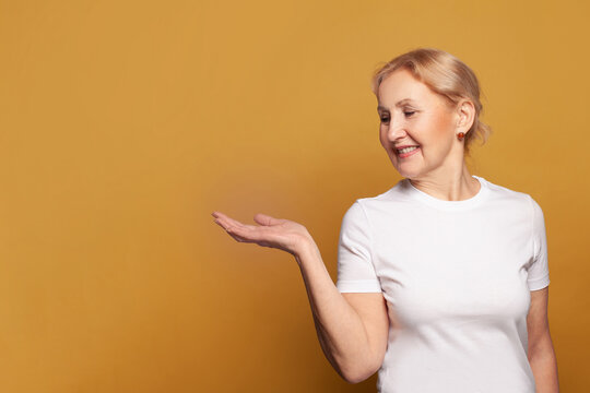 Mature Woman In White T-shirt With Empty Open Hand On Bright Yellow Studio Wall Background