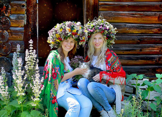 Two girls in large flower wreaths and national scarves on their shoulders are sitting with a cat. Background rural wooden house. Nature. Summer concept.