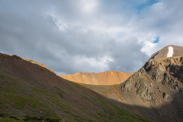 Dramatic alpine landscape with sunlit wide sharp mountain ridge under overcast sky at changeable weather. Atmospheric mountain scenery with large sharp rocks on ridge top in sunlight under cloudy sky.