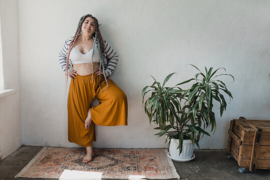 Positive smiling plus size woman with dreadlocks in bright yellow pants in a modern bohemian boho interior is meditating surrounded by large flowers and plants. Ecological approach to conscious consum