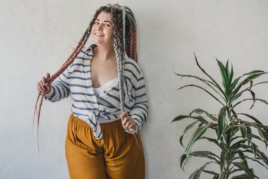 Positive smiling plus size woman with dreadlocks in bright yellow pants in a modern bohemian boho interior is meditating surrounded by large flowers and plants. Ecological approach to conscious consum