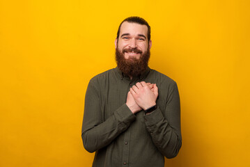 Young bearded man holding hands to heart and thinking about someone dear. Studio shot over yellow...