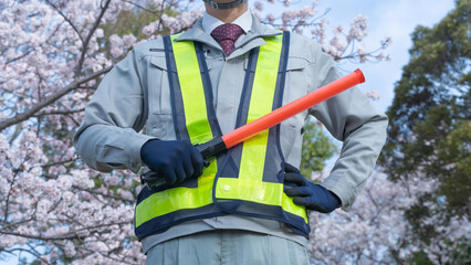 桜の背景で作業服で誘導棒を持つ人｜建設業・警備業