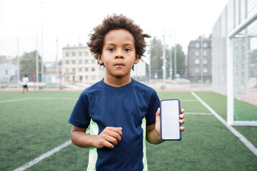 Kid using phone. Smiling and excited boy showing smartphone empty screen. Looking to camera. Cellphone display mockup mobile app advertisement. African american kid in football field. Blank screen