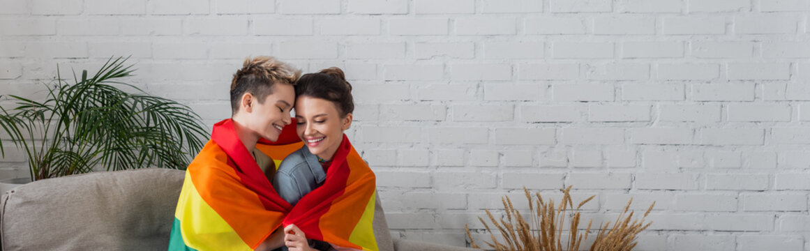 smiling pangender partners covered with lgbt flag near decorative plants at home, banner.
