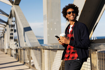 Smiling black man holding coffee to go. Happy African man using the phone outside..
