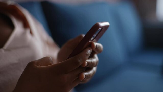 Close-up cropped shot of unrecognizable African-American woman sitting on blue sofa using typing on smartphone in modern room. Closeup hands of black female watching social media, scrolling content.